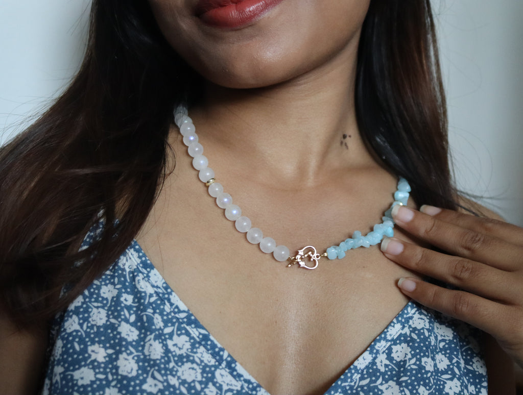 Woman wearing a light blue beaded necklace with a floral dress against a neutral background
