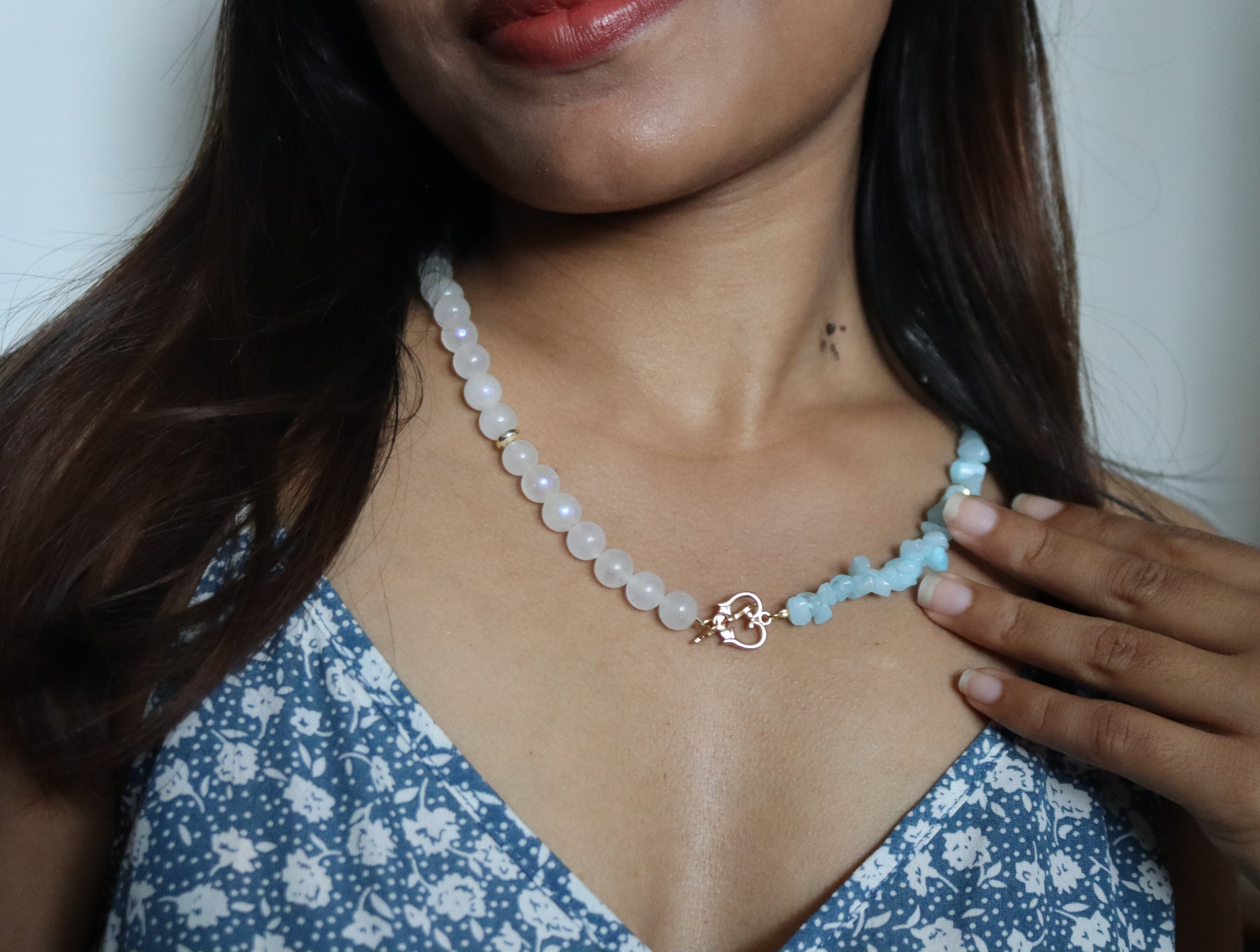 Woman wearing a light blue beaded necklace with a floral dress against a neutral background
